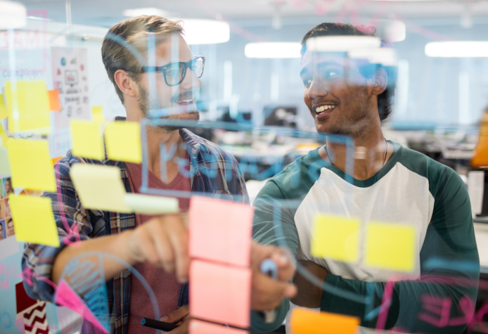 Two workers discussing work items shown on a visual Kanban board